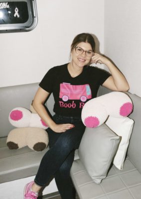 Rena Vanzo sits on a couch with pink and brown pillows shaped like breasts.