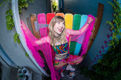 Stevie Shock in filmy pink sleeves in front of a rainbow picket fence.