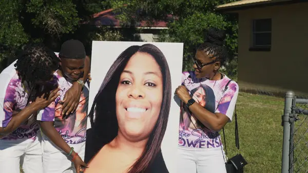 Three people hold up a poster of Ajike Owens in The Perfect Neighbor, directed by Geeta Gandbhir