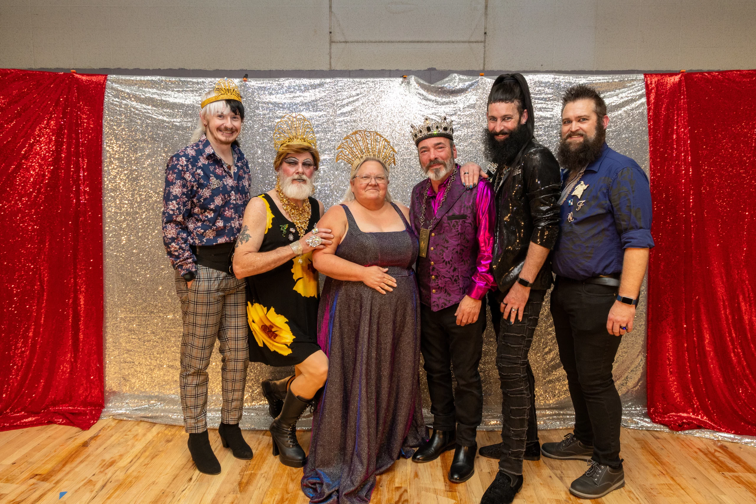 Six people dressed in elaborate outfits pose in front of a silver backdrop.