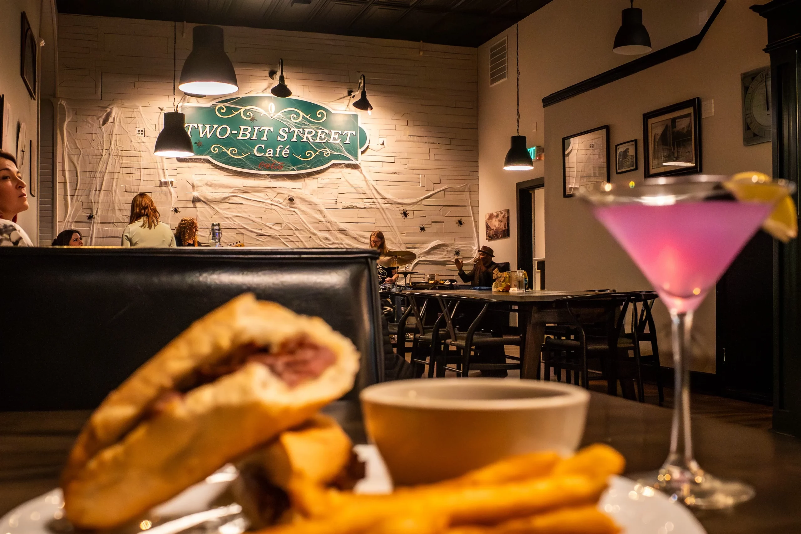A sandwich and a cocktail sit on a table at Two-Bit Bistro in Ogden, with the restaurant logo decorating the wall in the background.